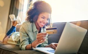 Smiling, young lady shops online with her debit card on her couch with her laptop. Get a debit card with your checking account!