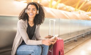 Young lady sits next to her luggage, with her phone in hand, listening to music. She is ready to travel with online and mobile banking.