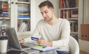 Young man sitting in front of his laptop, shopping with a prepaid credit card.