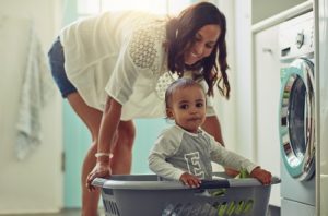 Young mother does laundry while her toddler sits in the laundry basket.